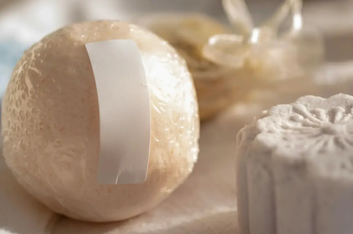 Close-up of handmade bath bombs with a blank white label on a round golden-wrapped one, alongside a decorative molded bath bomb in soft natural tones.