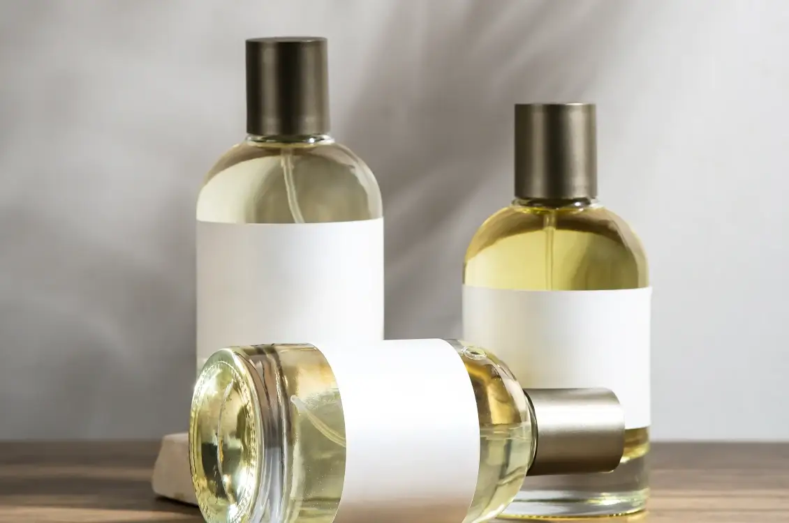 Three glass perfume bottles with dark caps and blank white labels, arranged on a light wooden surface against a soft gray background