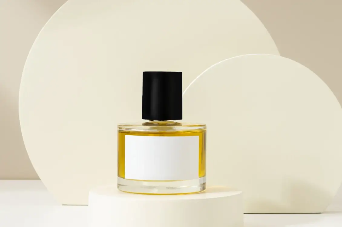 Glass perfume bottle with a black cap with a blank white label, displayed on a white pedestal against a minimalist beige backdrop.