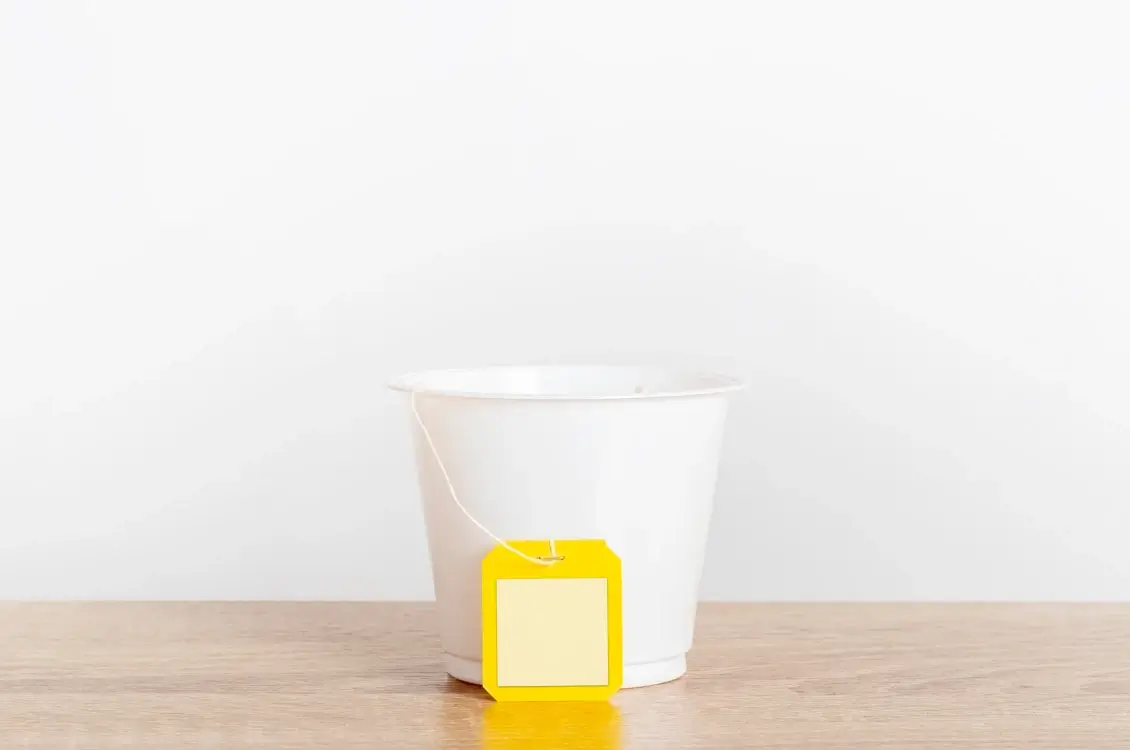 White tea cup on a light wooden surface with a yellow tea bag tag hanging over the side against a white background.