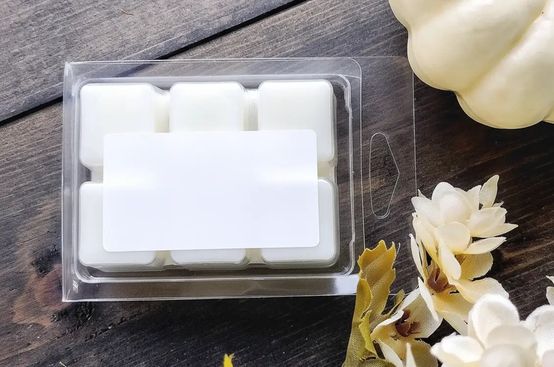White wax melt clamshell pack on a gray wooden surface next to a white pumpkin and white flowers with autumn leaves.